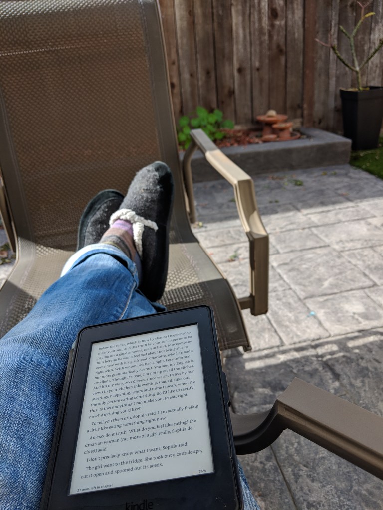 Feet clad in gray slippers crossed and propped on a brown chair on a sunny patio. The legs are in blue jeans and a Kindle Paperwhite is resting on them..