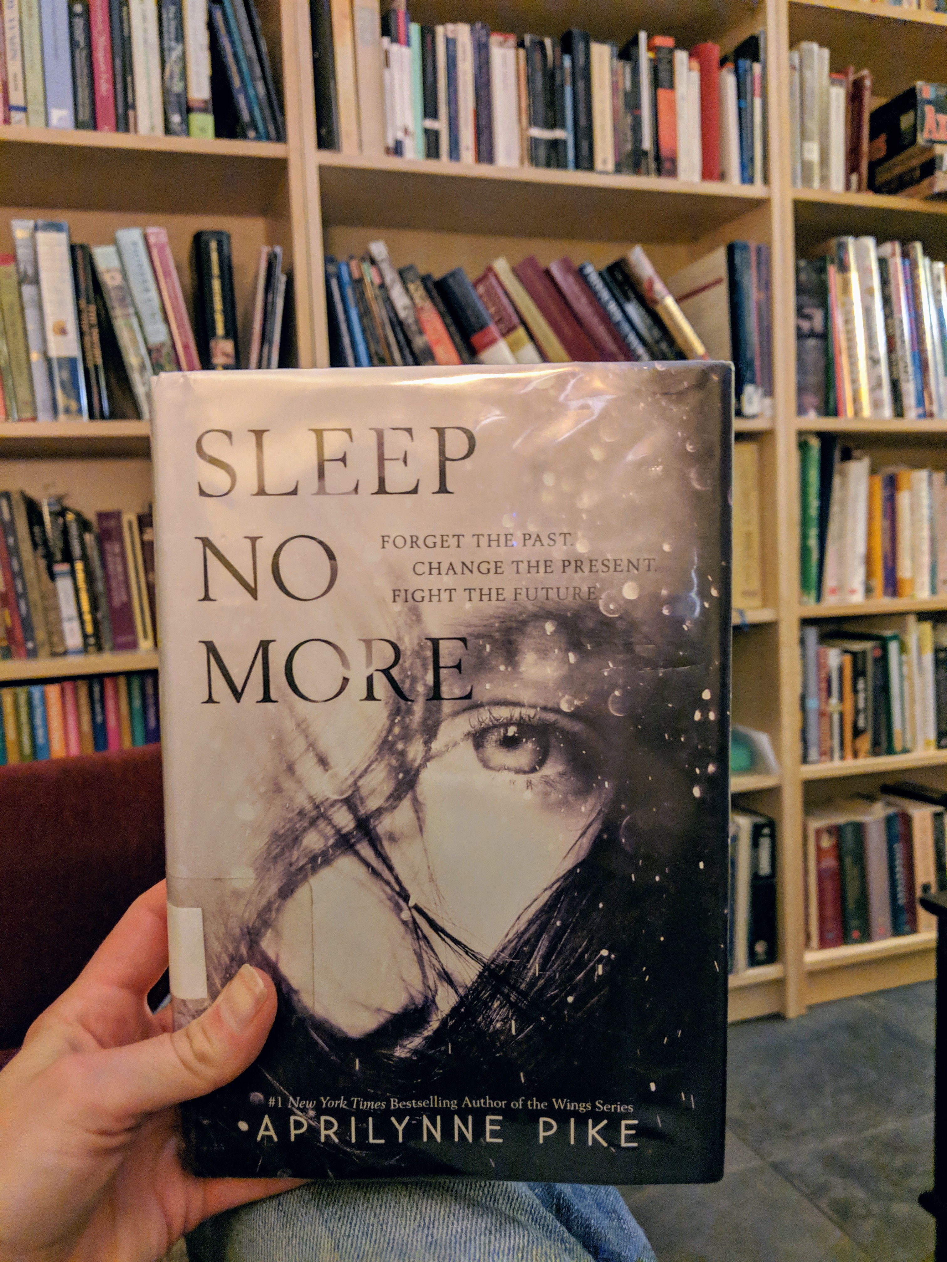 A hand holding a book in front of a bookshelf. The cover of the book has a black-and-white image of a woman's face overlaid with the title Sleep No More and the words Forget the Past, Change the Present, Fight the Future. Aprilynne Pike is printed across the bottom of the book cover.